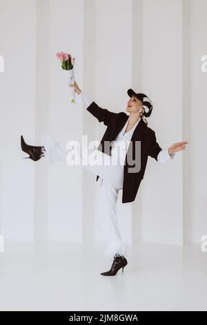 Eine fröhliche, stilvolle Frau in einem modischen Outfit, ein hellweißer Anzug mit Federn, eine schwarze Jacke, eine Baseballmütze und mit einem Blumenstrauß in ihrem h Stockfoto