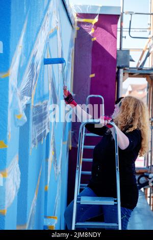 Erwachsene reife Rotschopf Malerin Frau mit lockigen roten Haaren und Sonnenbrille, steht auf Leiter und malt künstlerischen polygonalen Farbfleck in blauer Farbe Stockfoto