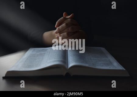 Frau reicht die Bibel an. Sie liest und betet über die bibel in einem dunklen Raum über einem Holztisch. Stockfoto