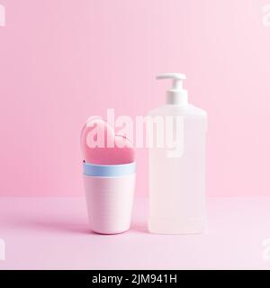 Quadratische Zusammensetzung mit Spülmitteln. Weiße Flasche Spülmittel und rosa Herzputzschwamm in sauberen Plastikbechern auf pastellrosa. Stockfoto