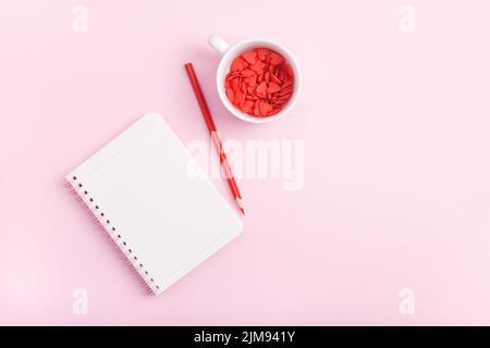 Leere Notizbuchseite, Bleistift und Becher voller roter, süßer Herzen auf pastellrosa Hintergrund. Valentinstag-Konzept. Stockfoto