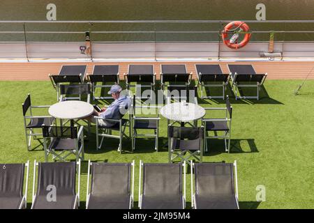 Mann, der auf dem Deck eines Ausflugsboots am Rheinufer sitzt, Köln, Deutschland. Mann sitzt auf dem Deck eines Ausflugsschiefes am Rheinuf Stockfoto