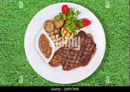 Gegrilltes Steak, gebackene Kartoffeln und Gemüse auf weißem Teller auf grünem Gras. Stockfoto