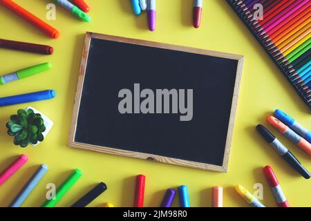 Kreidetafel mit farbigen Markern und Bleistiften auf gelbem Hintergrund. Schulbedarf für Kinder. Draufsicht. Stockfoto