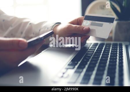 Weibliche Hände mit Kreditkarte und Telefon in der Nähe des Laptops. Nahaufnahme. Online-Zahlungen. Banküberweisung. Stockfoto
