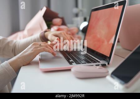 Weibliche Hände mit Laptop. Weibliche Büro Schreibtisch Arbeitsplatz Homeoffice Nachbau mit Laptop, rosa Tulpenblüten Blumenstrauß, Smartphone und rosa Accessoires. Stockfoto