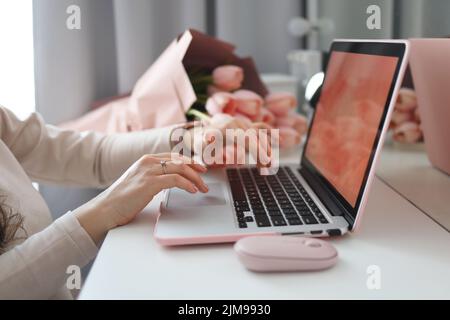 Weibliche Hände mit Laptop. Weibliche Büro Schreibtisch Arbeitsplatz Homeoffice Nachbau mit Laptop, rosa Tulpenblüten Blumenstrauß, Smartphone und rosa Accessoires. Stockfoto