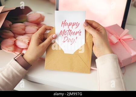 Die Hände einer Frau halten einen Brief in einem handgefertigen Umschlag. Rosa Hintergrund, Muttertagskonzept. Tulpen Blume und rosa Geschenkbox im Hintergrund. Damen Home de Stockfoto