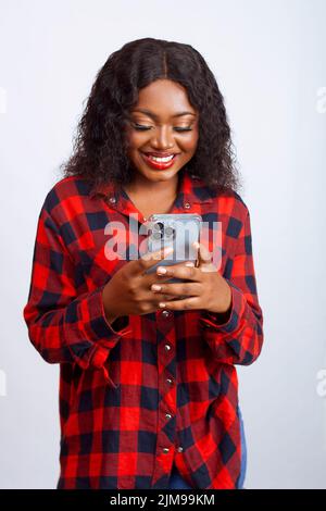 Schöne afrikanische Dame, die ihr Smartphone benutzt, das überrascht und aufgeregt aussieht, während sie Inhalte auf ihrem Telefon ansieht – isolierter weißer Hintergrund Stockfoto