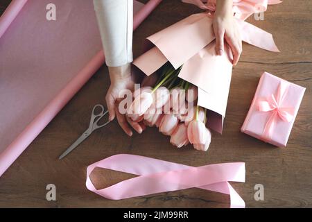 Weibliche Hände wickeln einen Haufen rosa Tulpen in rosa Kraftpapier neben einer Schere und einem Jute-Seil auf einen Holztisch, Draufsicht. Flache Lay-Zusammensetzung von mak Stockfoto