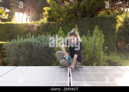 Junge Frau, die an einer Solarpaneelanlage arbeitet, um erneuerbare Energie mit grünem Garten im Hintergrund zu erhalten Stockfoto