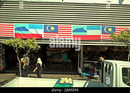 Menschen, die auf einem Gehweg in einem Hintergrund von allgemeinen Geschäften gehen, wo die Flagge von Sabah und die Nationalflagge Malaysias in Kota Kinabalu, Sabah, Malaysia, zu sehen sind. Stockfoto