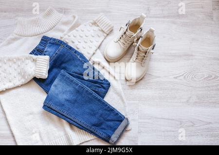 Damen flache Herbstkleidung mit warmweißem Pullover, blauen Jeans und weißen Schuhen, feminin modisches Outfit auf weißem Laken, Copy Space Stockfoto