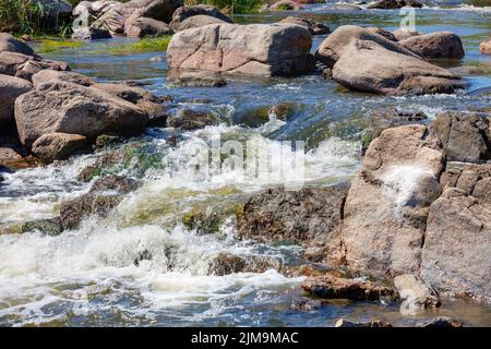 An einem sonnigen Sommertag sät zwischen Felsblöcken ein blauer Wasserstrom. Stockfoto