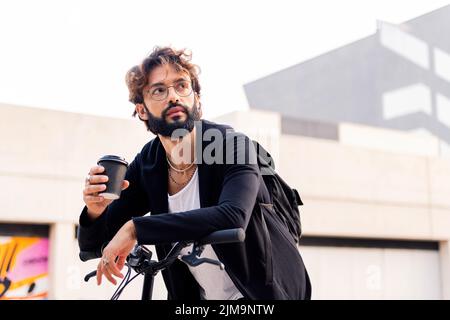 Der junge Kaukasusmann macht eine Pause, während er auf seinem Fahrrad einen Kaffee zum Mitnehmen trinkt, urbanes Lifestyle-Konzept und nachhaltige Mobilität, Kopierraum Stockfoto