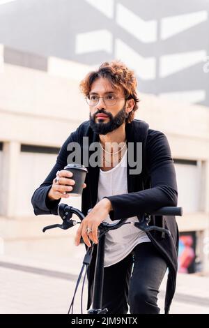 Der junge Kaukasusmann macht eine Pause, während er auf seinem Fahrrad einen Kaffee zum Mitnehmen trinkt, ein urbanes Lifestyle-Konzept und nachhaltige Mobilität Stockfoto