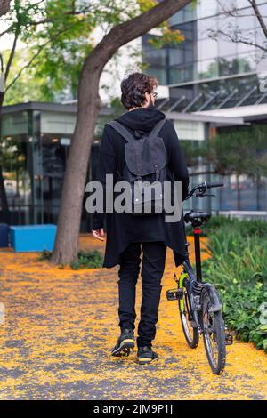 Rückansicht eines nicht erkennbaren Studenten, der mit einem Faltrad im Park des Universitätscampus läuft, Konzept von Jugend und Bildung Stockfoto