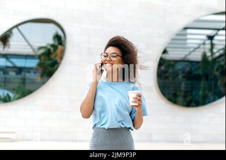 Telefongespräch. Eine geschäftige, freundliche afroamerikanische Frau mit lockigen Haaren, in formeller Kleidung, mit Brille, die in der Nähe des Business Centers im Freien läuft, eine Tasse Kaffee zum Mitnehmen hält, mit dem Smartphone spricht und lächelt Stockfoto