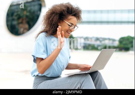 Virtuelles Meeting-Konzept. Beschäftigt erfolgreiche afroamerikanische junge Frau, Geschäftsfrau, sitzt im Freien, hält Online-Videokonferenz mit Laptop, diskutieren Finanzstrategie mit Kollegen, lächelnd Stockfoto