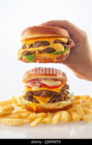 Hand hält einen Gourmet-Käse-Burger auf einem anderen Rindfleisch-Burger mit Kartoffelchips auf dem Boden Stockfoto