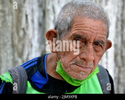 Der arme alte mexikanische Rentner mit selbstgemachter Gesichtsmaske aus grünem Stoff unter seinem Kinn blickt den Zuschauer lautlos an. Stockfoto