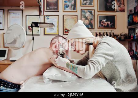 Junge blonde Frau Meister Tätowierer in Gläser und Hut mit Tattoo-Maschine von Hand machen tattoo-Dreamcatcher zur hand des Menschen. Stockfoto