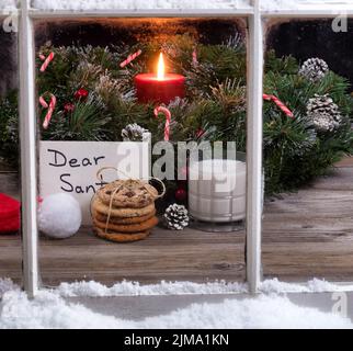 Verschneite Fensteransicht von Keksen und Milch plus Karte für den Weihnachtsmann mit brennender Kerze im Hintergrund Stockfoto