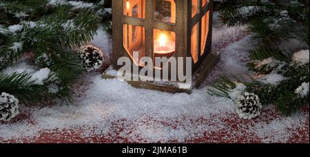 Vintage Laterne mit brennender Kerze auf verschneiten rustikalen roten Holzbrettern mit Weihnachtsdekorationen Stockfoto