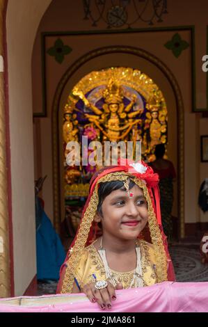 Howrah, Indien -26.. Oktober 2020 : Bengalisches Mädchen in festlichem Kleid, lächelnd und posiert mit Göttin Durga im Hintergrund, im alten Haus. Stockfoto