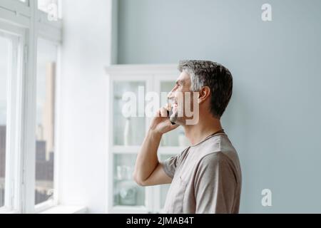 Lächelnder Mann, der auf einem Smartphone neben dem Fenster steht. Stockfoto