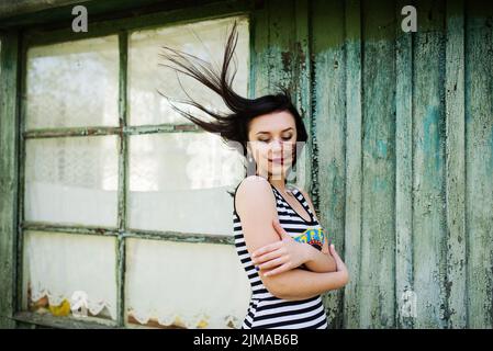 Brünette Modell Mädchen mit Haaren auf der Luft am Kleid mit Streifen Hintergrund Cian Holzhaus mit alten Fenstern zu tragen. Stockfoto