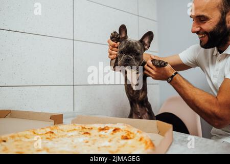 Fröhlicher Mann mit französischer Bulldogge von Paws vor der Pizza am Küchentisch, kleiner Hund wartet ungeduldig auf menschliches Essen Stockfoto