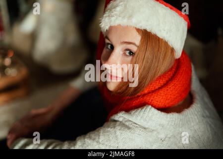 Fröhliche kaukasische weibliche Teenager in Weihnachtsmannhut und rotem Schal mit Neujahrsdekoration und schwachem Licht. Junges Mädchen feiert Neujahr 2023. Hochwertige Fotos Stockfoto