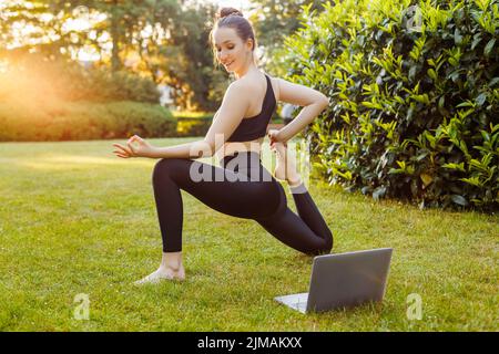 Junge Frau mit Laptop praktiziert Yoga im Sommerpark. Konzept der Outdoor Online-Yoga-Kurse, Fitness-Lernen oder Unterricht, aktive und sportliche lif Stockfoto