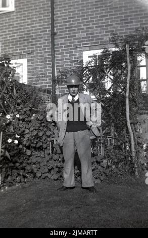 1950s, ein älterer Mann, der in einem Hintergarten steht, Jacke und Krawatte sowie Pullover, mit einem alten Metallkampfhelm auf dem Kopf, England, Großbritannien. Ein Stahl-Kampfhelm, bekannt als der "Brodie-Helm", nach seinem Schöpfer John Leopold Brodie, der sein ursprüngliches Design im Jahr 1915 patentieren ließ, wurden modifizierte Versionen bekannt als: Battle Bowler, tommy Hut und Zinnhut. Metallkampfhüte wurden kurz nach Beginn des Jahres WW1 geschaffen, da die Kopfbedeckungen aus Stoff und Leder, die traditionell von den Soldaten getragen wurden, keinen Schutz vor Artillerie boten. Der Helm wurde auch WW2 getragen, ebenso von Polizei, Feuerwehr und ARP-Aufsehern. Stockfoto