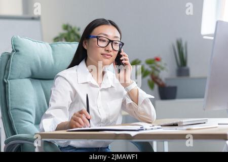 Porträt einer jungen, schönen asiatischen Geschäftsfrau in einer Brille in einem weißen Hemd, die am Telefon spricht. Er sitzt am Tisch im Büro und hält eine kleine Hand. Stockfoto