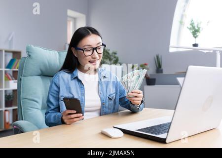 Business-Training über Geld. Eine junge asiatische Geschäftsfrau führt eine Schulung zum Thema Geld durch. Er hält ein Telefon und eine Schnur von Rechnungen. Sitzt an seinem Schreibtisch, redet, schaut in die Kamera. Stockfoto