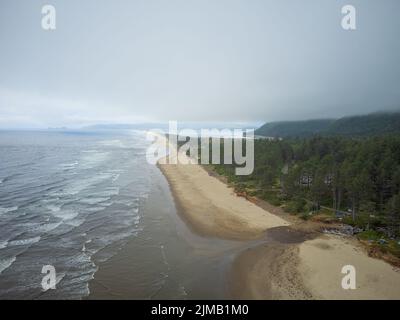 Schießen von einer Drohne. Kleine gemütliche Häuser an der grünen Hügelküste des Ozeans. In der Ferne ist eine Bergkette sichtbar. Lichtwellen des Ozeans Stockfoto