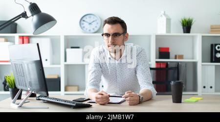 Ein junger Mann in Gläsern steht in der Nähe eine Tabelle im Büro und hält einen Bleistift in der Hand. Stockfoto