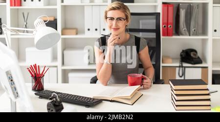 Ein junges Mädchen sitzt an einem Tisch im Büro, mit einem Bleistift und einem roten Becher in der Hand. Vor dem Mädchen liegt ein offenes Buch. Stockfoto