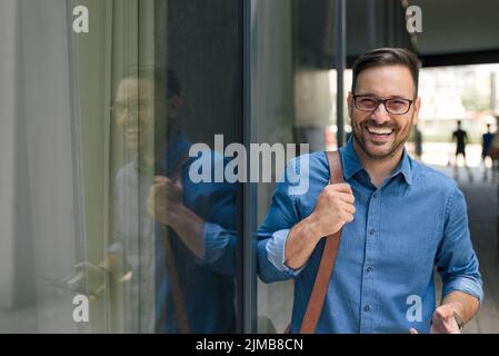 Porträt eines lächelnden jungen professionellen Laptop-Tasche. Der Geschäftsmann trägt formelle Formen. Er steht am Fenster des Bürogebäudes in der Stadt. Stockfoto