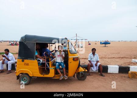 Ein Junge, der in einem touk touk sitzt, lächelt zur Kamera, während andere Leute sich am Marina Beach in Chennai amüsieren. Stockfoto