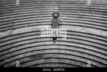 Eine graue Ansicht von oben einer Frau auf ihren Knien, die vor einer Statue auf einer Treppe betet Stockfoto