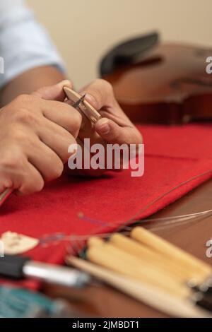 Vertikales Foto der Hände eines Arbeiters, der in einer Werkstatt mit einem kleinen Messer eine Geigenbrücke formt Stockfoto