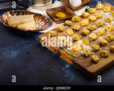 Der Prozess der Herstellung von hausgemachten Knödeln. Appetitliche Knödel auf einem Holzschneidebrett und Zutaten auf dunkelblauem Hintergrund. Low-Angle-Ansicht. Stockfoto