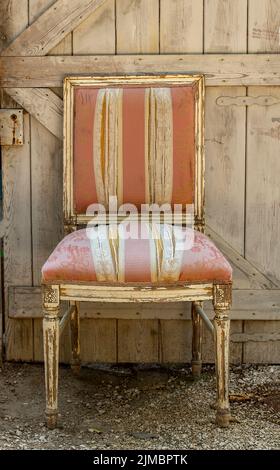 Shabby chic gepolsterten einzigen antiken Stuhl mit verblassten Farbe und links außerhalb gegen eine rustikale Holztür. Alte Stuhl links zu verfaulen, Shabby chic Stil. Stockfoto