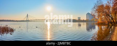 Panoramablick auf das Ufer des Dnjepr-Flusses mit Herbstbäumen und der Pivnichnyj-Brücke am Horizont, Kiew, Ukraine Stockfoto