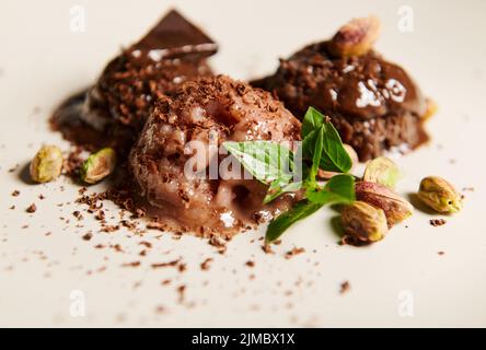 Selektive Konzentration auf Kugeln Schokoladeneis, gekrönt mit Pistazien- und Zitronenbasilblättern, die auf einem weißen Teller schmelzen Stockfoto