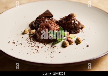 Nahaufnahme. Stillleben. Gefrorene Kugeln aus Schokoladeneis, mit Pistazien- und Zitronenbasilblättern, schmelzen auf weißem Teller Stockfoto