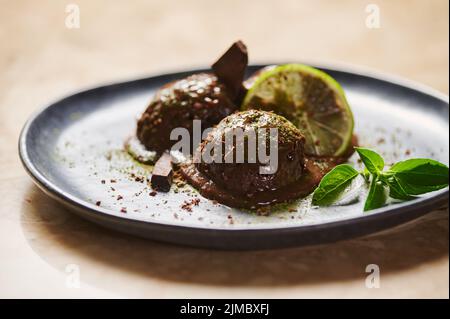 Kugeln Schokoladensorbet, mit bestreutem Matcha-Pulver, Limettenscheibe, Zitronenbasil auf dunkelblauem Teller. Selektiver Fokus Stockfoto